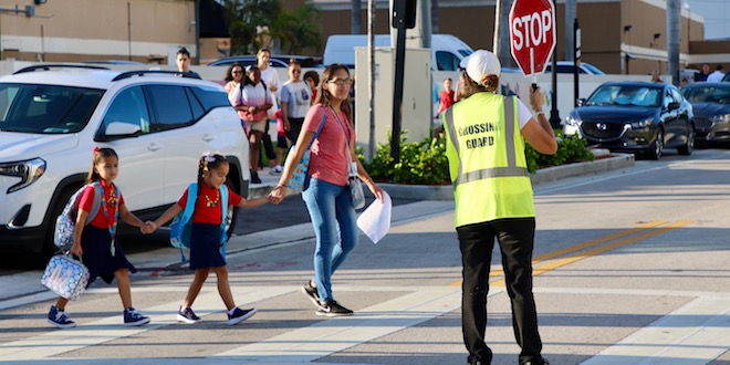 Mother and her two children cross the street on the way to school with crossing guard holding a stop sign for traffic.