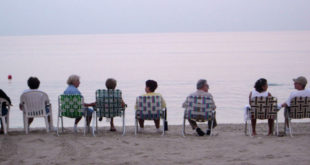 Seniors sitting in a row of lawn chairs on the beach facing the ocean.